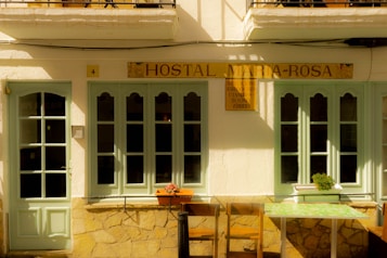 A quaint facade of a small hostal featuring light green doors and windows set in a cream-colored wall. The sign above reads 'Hostal Maria-Rosa'. There are two chairs and a small table with a floral pattern in front, and a potted plant sits on the windowsill.