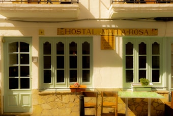 A quaint facade of a small hostal featuring light green doors and windows set in a cream-colored wall. The sign above reads 'Hostal Maria-Rosa'. There are two chairs and a small table with a floral pattern in front, and a potted plant sits on the windowsill.