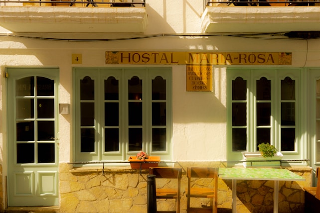 A quaint facade of a small hostal featuring light green doors and windows set in a cream-colored wall. The sign above reads 'Hostal Maria-Rosa'. There are two chairs and a small table with a floral pattern in front, and a potted plant sits on the windowsill.