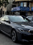 A sleek, modern car parked in an urban environment with reflective glass windows on surrounding buildings. Above, signage in multiple languages includes the name of a bank. The car is metallic gray with a sharp design and intricate alloy wheels.