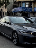 A sleek, modern car parked in an urban environment with reflective glass windows on surrounding buildings. Above, signage in multiple languages includes the name of a bank. The car is metallic gray with a sharp design and intricate alloy wheels.
