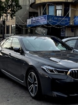 A sleek, modern car parked in an urban environment with reflective glass windows on surrounding buildings. Above, signage in multiple languages includes the name of a bank. The car is metallic gray with a sharp design and intricate alloy wheels.