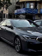 A sleek, modern car parked in an urban environment with reflective glass windows on surrounding buildings. Above, signage in multiple languages includes the name of a bank. The car is metallic gray with a sharp design and intricate alloy wheels.