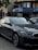 A sleek, modern car parked in an urban environment with reflective glass windows on surrounding buildings. Above, signage in multiple languages includes the name of a bank. The car is metallic gray with a sharp design and intricate alloy wheels.