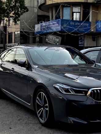 A sleek, modern car parked in an urban environment with reflective glass windows on surrounding buildings. Above, signage in multiple languages includes the name of a bank. The car is metallic gray with a sharp design and intricate alloy wheels.