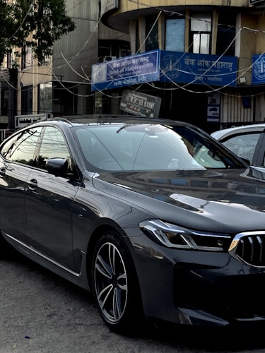 A sleek, modern car parked in an urban environment with reflective glass windows on surrounding buildings. Above, signage in multiple languages includes the name of a bank. The car is metallic gray with a sharp design and intricate alloy wheels.