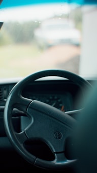 The image focuses on the interior of a vehicle, highlighting the steering wheel with a Volkswagen logo. Behind the wheel, the dashboard with gauges is slightly visible. The background is blurred, showing a glimpse of a grassy area and an ambiguous vehicle in the distance.