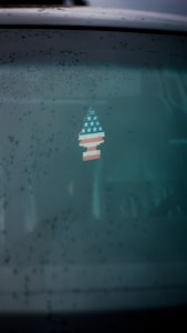 A rearview mirror displays a dangling air freshener shaped like a tree with an American flag design. The window is covered with raindrops and a slight foggy effect.