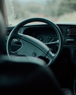 A close-up view of the interior of a car, focusing on a steering wheel with the Volkswagen logo in the center. The dashboard in the background features speedometer gauges, and there is a hint of sunlight filtering through the glass, casting soft reflections.