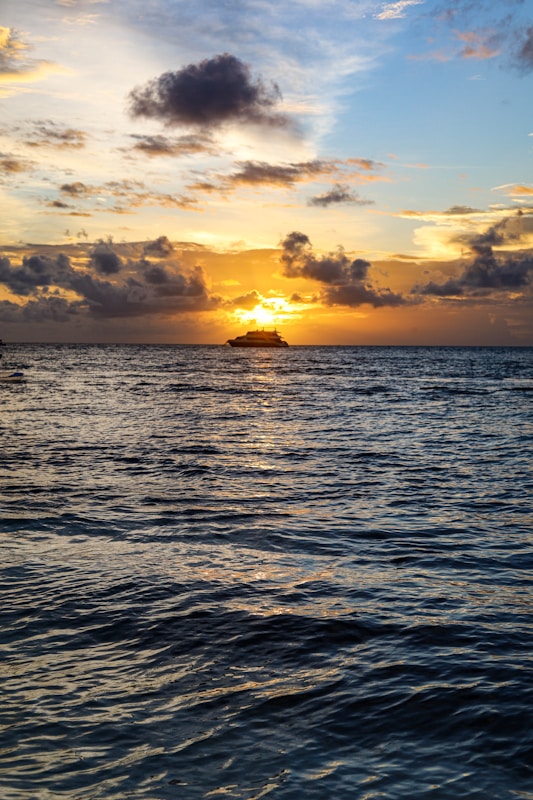 A serene ocean scene at sunset, with a silhouette of a cruise ship on the horizon. The sky is filled with a mix of dark and golden clouds, reflecting the warm tones on the rippling water surface.