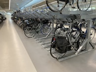 A spacious indoor bicycle parking area with multiple rows of bikes neatly arranged on a two-tiered rack system. The area is well-lit, featuring polished concrete floors and a modern design with metal framework supporting the bicycles. Some bikes have baskets or panniers attached.