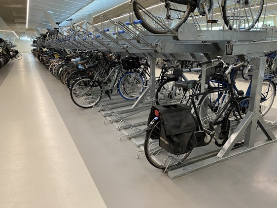 A spacious indoor bicycle parking area with multiple rows of bikes neatly arranged on a two-tiered rack system. The area is well-lit, featuring polished concrete floors and a modern design with metal framework supporting the bicycles. Some bikes have baskets or panniers attached.