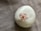 A smooth, glossy white ceramic object is placed on a textured fabric surface. It features a small, ornamental flower with pale pink petals and a red center at its top.