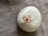 A smooth, glossy white ceramic object is placed on a textured fabric surface. It features a small, ornamental flower with pale pink petals and a red center at its top.