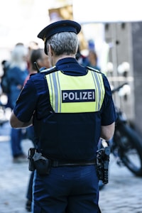 A police officer is seen from behind, wearing a dark blue uniform and a high-visibility vest with the word 'POLIZEI' written on it. The officer is in a public setting with several people around, some of whom appear blurred in the background, suggesting movement or activity. There is a bicycle nearby and the scene is lit by daylight.