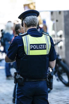 A police officer is seen from behind, wearing a dark blue uniform and a high-visibility vest with the word 'POLIZEI' written on it. The officer is in a public setting with several people around, some of whom appear blurred in the background, suggesting movement or activity. There is a bicycle nearby and the scene is lit by daylight.