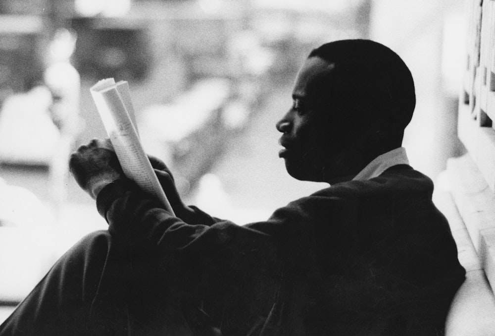 A man, from the side, sitting against a wall and reading a newspaper.