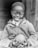 A young child with a joyful smile holds a round object in both hands. Dressed in a denim overall and a hoodie, the child exudes a sense of warmth and happiness. The background is out of focus, emphasizing the child's expression and attire.