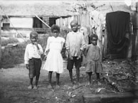 Four young children stand barefoot on a dirt ground in a rural setting. The children are dressed in simple, worn clothing. Wooden fencing and a rustic building are visible in the background, with some clothes hanging on the fence.
