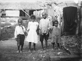 Four young children stand barefoot on a dirt ground in a rural setting. The children are dressed in simple, worn clothing. Wooden fencing and a rustic building are visible in the background, with some clothes hanging on the fence.