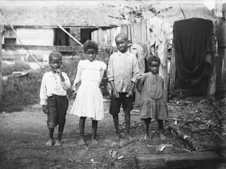 Four young children stand barefoot on a dirt ground in a rural setting. The children are dressed in simple, worn clothing. Wooden fencing and a rustic building are visible in the background, with some clothes hanging on the fence.