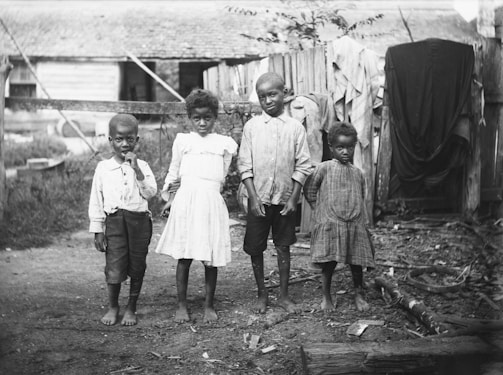 Four young children stand barefoot on a dirt ground in a rural setting. The children are dressed in simple, worn clothing. Wooden fencing and a rustic building are visible in the background, with some clothes hanging on the fence.