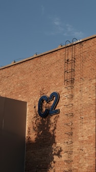 A brick wall with a metal ladder attached vertically. The letters 'C2' are prominently displayed in large blue font. Shadows from nearby trees are cast on the wall, with a clear blue sky above.