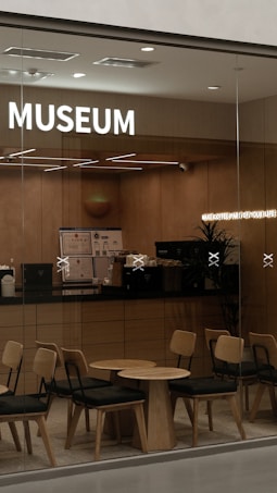 A modern caf&eacute; setting with wooden furniture, including chairs and tables arranged neatly. The counter area is visible with a coffee machine and other caf&eacute; essentials. Soft lighting creates a warm, inviting atmosphere. The word 'MUSEUM' is prominently displayed on the wall, and there's a secondary illuminated sign that reads 'MAKE COFFEE PART OF YOUR LIFE'.