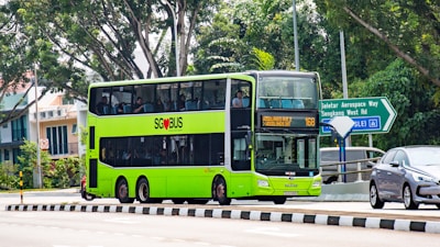 A bright green double-decker bus with 'SG BUS' logo is traveling on a street surrounded by lush green trees. Nearby, a road sign points towards Seletar Aerospace Way and Sengkang West Rd. A car follows the bus on the road, and the bus is displaying its route number as 168.