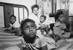 An African American woman and several children sitting on a bed in an apartment.