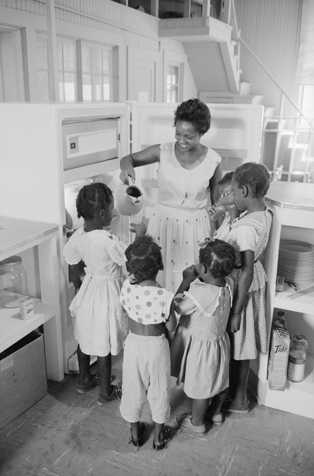 African American woman at refrigerator holding a pitcher as she pours out drinks for children, probably Little Rock, Arkansas.