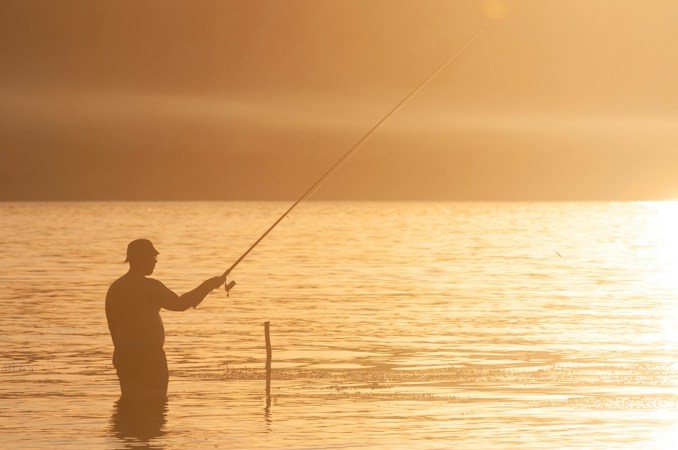 Silhouette of an angler in a cap standing waist-deep in a lake, casting a long rod into golden evening light