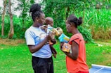 Two women stand outdoors with lush greenery in the background. One woman holds a baby and offers a packet of goods to the other woman. They appear to be engaged in an act of sharing or distribution.