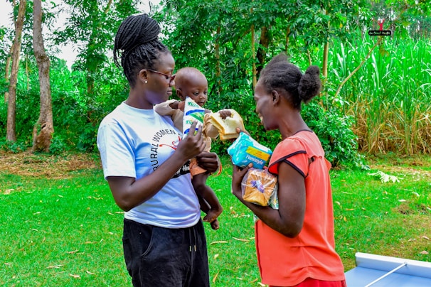 Two women stand outdoors with lush greenery in the background. One woman holds a baby and offers a packet of goods to the other woman. They appear to be engaged in an act of sharing or distribution.