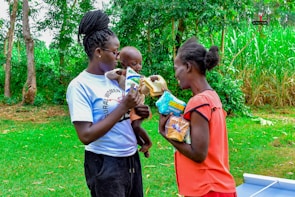Two women stand outdoors with lush greenery in the background. One woman holds a baby and offers a packet of goods to the other woman. They appear to be engaged in an act of sharing or distribution.