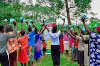 A group of women and children stand outdoors holding sanitary products above their heads. They form a semi-circle in a lush green environment with tall trees and grass surrounding them. A banner with text and logos appears in the background.