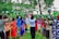 A group of women and children stand outdoors holding sanitary products above their heads. They form a semi-circle in a lush green environment with tall trees and grass surrounding them. A banner with text and logos appears in the background.