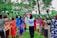 A group of women and children stand outdoors holding sanitary products above their heads. They form a semi-circle in a lush green environment with tall trees and grass surrounding them. A banner with text and logos appears in the background.