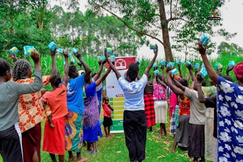 A group of women and children stand outdoors holding sanitary products above their heads. They form a semi-circle in a lush green environment with tall trees and grass surrounding them. A banner with text and logos appears in the background.