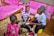 Three individuals are gathered on the floor handling maize in a room. The room has wooden furniture covered with pink fabric. One person is holding a maize cob while the others are engaging in conversation. A black container filled with more maize cobs is present, and packets of products are on the furniture.