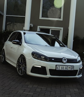 A white Volkswagen car is parked on a paved driveway in front of a modern building with large windows and white columns. The vehicle features sleek alloy wheels and a license plate with visible characters. There is a reflection of trees on the car and windows.