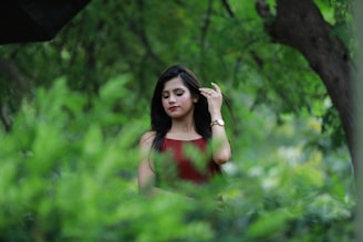 A young woman with long black hair is standing amidst lush green foliage in a natural setting. She is wearing a sleeveless red top and a wristwatch. Her hand is raised to adjust her hair, and she appears calm and serene.