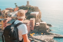 A person wearing a beige cap and a backpack gazes towards a scenic coastal town. The town features colorful buildings and a tower overlooking the sea. The atmosphere is relaxed with people and umbrellas by the waterfront.