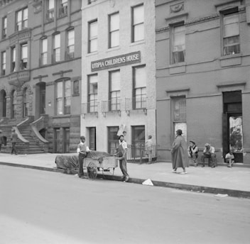 A black-and-white photograph depicts a city street with several brick buildings. Prominently displayed is a building with a sign reading 'Utopia Children's House'. The scene includes people going about their day; two individuals are standing by a cart on the street, and a group of people, including a child, are sitting and standing on the sidewalk. The architecture suggests an older urban setting, likely from the mid-20th century.