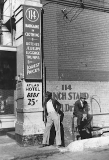 Two men are standing and sitting outside a brick building with a sign advertising bargains in clothing, watches, jewelry, luggage, trunks, lowest prices, and hotel beds for 25 cents. The clothing style suggests an older time period.
