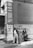 Two men are standing and sitting outside a brick building with a sign advertising bargains in clothing, watches, jewelry, luggage, trunks, lowest prices, and hotel beds for 25 cents. The clothing style suggests an older time period.