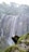 A person stands triumphantly on a large rock ledge in front of a tall, cascading waterfall. Lush greenery surrounds the area, and mist rises from the crashing water.