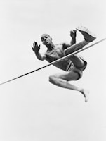 A male athlete is captured in mid-air performing a high jump. He is shirtless and wearing shorts, with a focused expression on his face. His body is contorted in an athletic pose, emphasizing his muscular build as he clears the bar.