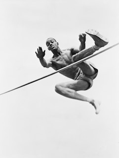 A male athlete is captured in mid-air performing a high jump. He is shirtless and wearing shorts, with a focused expression on his face. His body is contorted in an athletic pose, emphasizing his muscular build as he clears the bar.