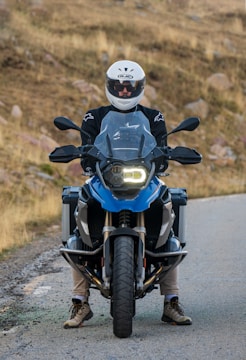 A person wearing a white motorcycle helmet and black jacket sits on a blue adventure motorcycle on a rural road. The background shows dry grass and rocky terrain.